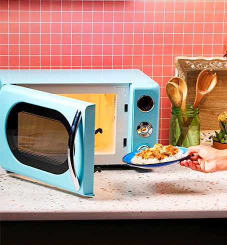 Person holding a plate of food in front of a blue microwave with a pink tiled wall background.