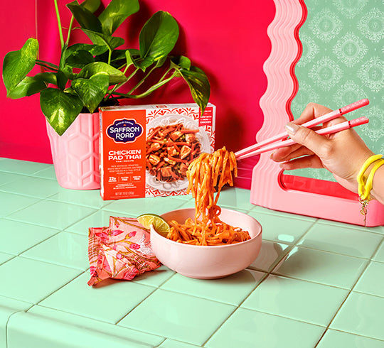 Person eating Saffron Road Chicken Pad Thai with pink chopsticks on a colorful background