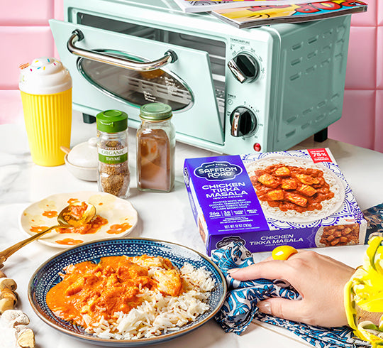 Mint green toaster oven on a kitchen counter with a plate of food, Saffron House Chicken Tikka Masala packaging, and a hand holding a cloth.