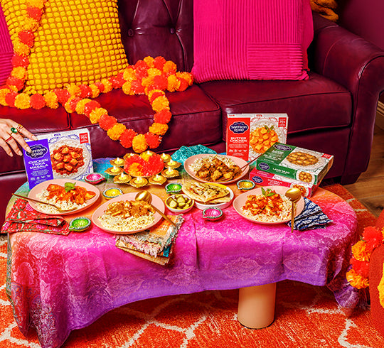 Table with food and snacks on a pink tablecloth, decorated with flowers and colorful pillows.